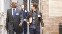 Dois homens andam lado a lado. Um homem negro de terno cinza, outro homem de jaqueta de couro. Ambos com crachás, num fundo urbano.