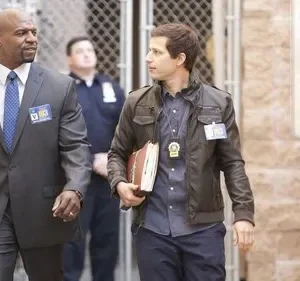 Dois homens andam lado a lado. Um homem negro de terno cinza, outro homem de jaqueta de couro. Ambos com crachás, num fundo urbano.