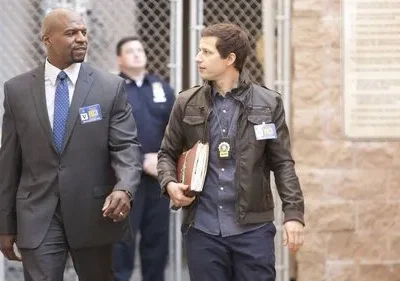 Dois homens andam lado a lado. Um homem negro de terno cinza, outro homem de jaqueta de couro. Ambos com crachás, num fundo urbano.