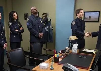 Seis policiais em uniforme azul numa sala. Dois homens sorriem ao apertar as mãos, enquanto quatro colegas os observam.