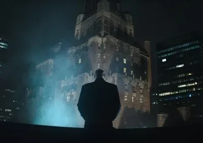 Homem em silhueta observa uma cidade noturna e nebulosa. Edifícios altos iluminados criam um clima misterioso.