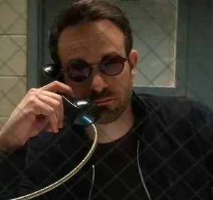 Homem barbudo de óculos escuros redondos fala ao telefone em cela. Uma grade quadriculada sobrepõe a imagem. Clima sério.