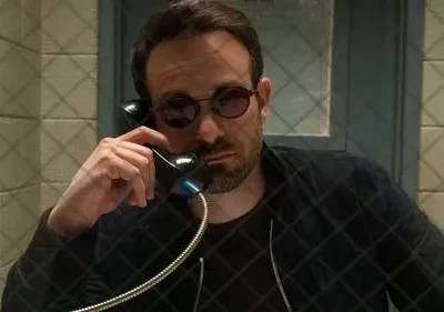 Homem barbudo de óculos escuros redondos fala ao telefone em cela. Uma grade quadriculada sobrepõe a imagem. Clima sério.