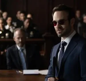 Homem de terno azul e óculos escuros sorri confiante em tribunal. Pessoas ao fundo observam, clima de drama legal.