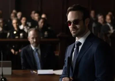 Homem de terno azul e óculos escuros sorri confiante em tribunal. Pessoas ao fundo observam, clima de drama legal.