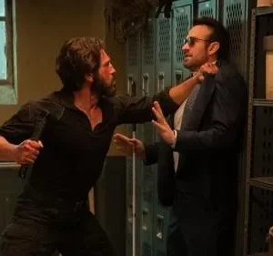 Dois homens lutam. Um barbudo, com machado, ataca outro de terno e óculos escuros em um vestiário escuro.