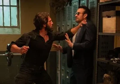 Dois homens lutam. Um barbudo, com machado, ataca outro de terno e óculos escuros em um vestiário escuro.