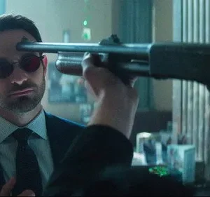 Homem de terno e óculos escuros, com ponto laser na testa, encara calmamente uma espingarda apontada por figura vermelha.