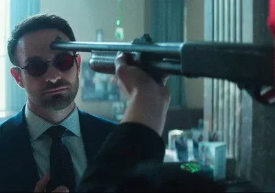 Homem de terno e óculos escuros, com ponto laser na testa, encara calmamente uma espingarda apontada por figura vermelha.