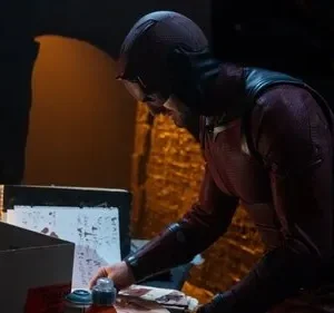 Personagem encapuzado em traje escuro com óculos examina papéis com manchas vermelhas em uma mesa. Ambiente escuro e sombrio.