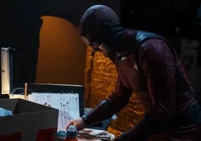 Personagem encapuzado em traje escuro com óculos examina papéis com manchas vermelhas em uma mesa. Ambiente escuro e sombrio.