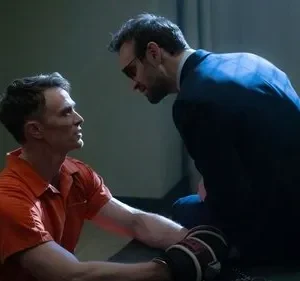 Um prisioneiro de macacão laranja olha intensamente para um homem de terno com óculos escuros, inclinados em uma sala sombria. Cena dramática.
