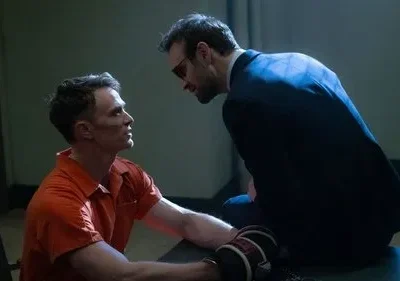 Um prisioneiro de macacão laranja olha intensamente para um homem de terno com óculos escuros, inclinados em uma sala sombria. Cena dramática.