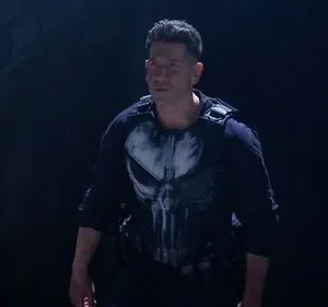 Homem sério, vestindo colete tático e camisa preta com caveira branca, em ambiente escuro e tenso.
