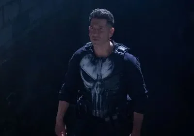 Homem sério, vestindo colete tático e camisa preta com caveira branca, em ambiente escuro e tenso.