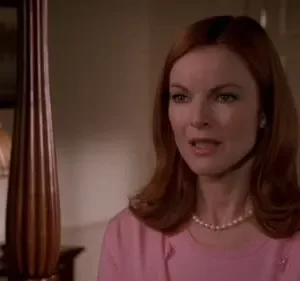 Mulher ruiva de blusa rosa e colar de pérolas com expressão atenta, em quarto de luz baixa. Cama com poste e luminária visíveis.