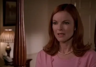 Mulher ruiva de blusa rosa e colar de pérolas com expressão atenta, em quarto de luz baixa. Cama com poste e luminária visíveis.