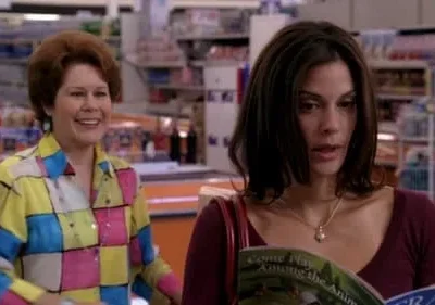 Duas mulheres em um mercado. Uma sorri com camisa colorida xadrez, a outra lê uma revista com expressão séria. Clima de cotidiano.