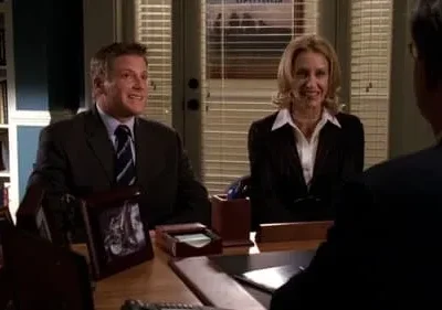 Dois executivos sorridentes, um homem e uma mulher, em uma mesa de escritório. Um terceiro de costas na frente. Ambiente profissional.