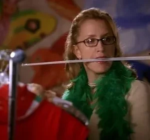 Mulher de óculos e boa verde equilibra uma espada decorativa na boca, com roupas em cabides e fundo colorido e abstrato.