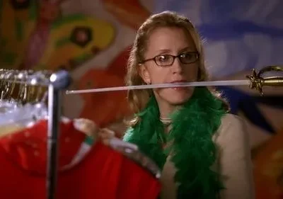 Mulher de óculos e boa verde equilibra uma espada decorativa na boca, com roupas em cabides e fundo colorido e abstrato.