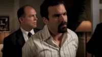 Dois homens em um ambiente interno. Um de barba em camisa listrada olha com preocupação. Outro, de terno, observa seriamente.