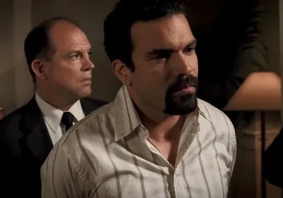 Dois homens em um ambiente interno. Um de barba em camisa listrada olha com preocupação. Outro, de terno, observa seriamente.