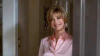 Mulher loira sorridente, com camisa rosa, espreita por trás de uma porta, com um ar amigável e acolhedor.