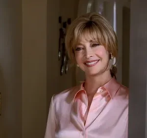 Mulher loira sorridente, com camisa rosa, espreita por trás de uma porta, com um ar amigável e acolhedor.