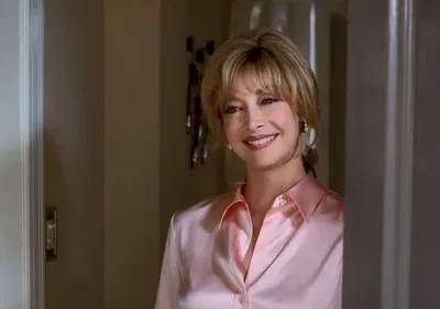 Mulher loira sorridente, com camisa rosa, espreita por trás de uma porta, com um ar amigável e acolhedor.
