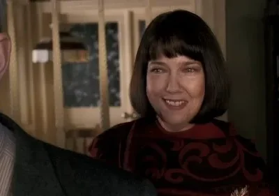 Mulher de bob escuro e blusa vermelha estampada sorri. Ombro de homem à esquerda. Cenário de casa acolhedora, luz suave.