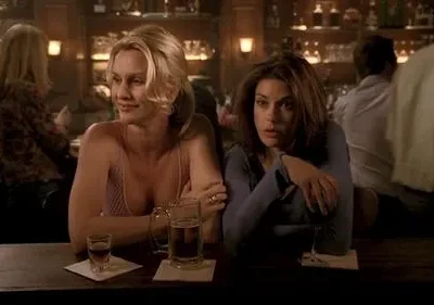 Duas mulheres em um bar. Uma loira sorri, a outra morena olha seriamente. Sentadas ao balcão com bebidas, ambiente intimista.