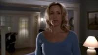 Uma mulher loira de suéter azul claro, com expressão séria, em uma sala com iluminação suave e detalhes domésticos.
