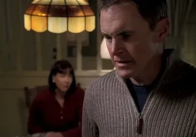 Homem em suéter bege, com expressão preocupada, olha para baixo. Mulher borrada o observa. Cena interna com luz quente.