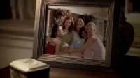 Retrato emoldurado: cinco mulheres sorridentes, de mãos dadas e se abraçando em frente a uma casa. Transmitem um clima de forte amizade.
