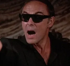 Um homem de óculos escuros e camisa preta grita, apontando diretamente para frente. O ambiente é escuro e dramático.