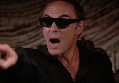 Um homem de óculos escuros e camisa preta grita, apontando diretamente para frente. O ambiente é escuro e dramático.