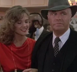 Homem de chapéu e terno sério, junto a mulher sorridente de vestido vermelho, em cenário elegante com luzes quentes.