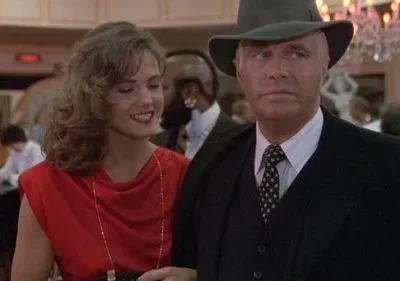 Homem de chapéu e terno sério, junto a mulher sorridente de vestido vermelho, em cenário elegante com luzes quentes.