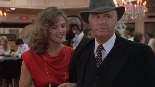 Homem de chapéu e terno sério,junto a mulher sorridente de vestido vermelho,em cenário elegante com luzes quentes.