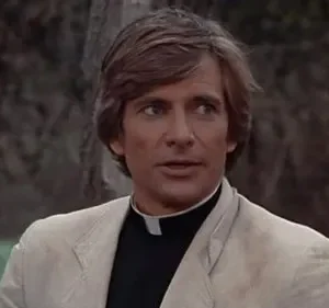 Homem de cabelo castanho, camisa clerical e casaco claro, olhando para o lado. Fundo desfocado de árvores e folhagem. Clima atento.