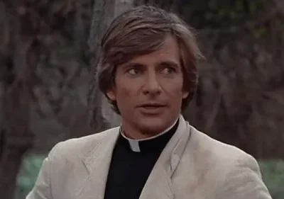 Homem de cabelo castanho, camisa clerical e casaco claro, olhando para o lado. Fundo desfocado de árvores e folhagem. Clima atento.