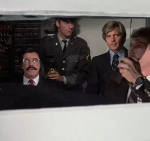 Cinco homens, incluindo um de uniforme e outro de bigode, olhando seriamente de dentro de uma cabine com painéis técnicos.