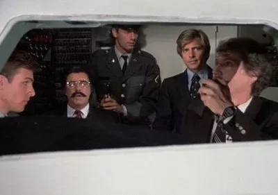 Cinco homens, incluindo um de uniforme e outro de bigode, olhando seriamente de dentro de uma cabine com painéis técnicos.