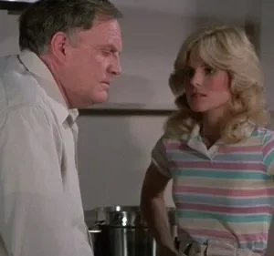 Um homem mais velho e uma mulher loira de blusa listrada. Ele parece pensativo; ela, séria e com as mãos na cintura.