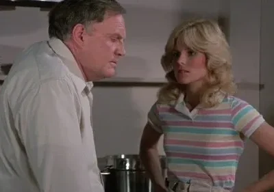 Um homem mais velho e uma mulher loira de blusa listrada. Ele parece pensativo; ela, séria e com as mãos na cintura.