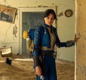 Mulher séria de uniforme azul, mochila, em casa em ruínas com areia no chão e paredes descascando.