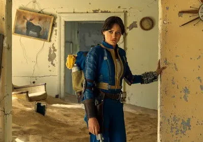 Mulher séria de uniforme azul, mochila, em casa em ruínas com areia no chão e paredes descascando.