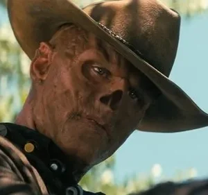 Close-up de um homem com chapéu de cowboy e rosto gravemente desfigurado, cicatrizado, com um olhar fixo e intenso sob céu azul.