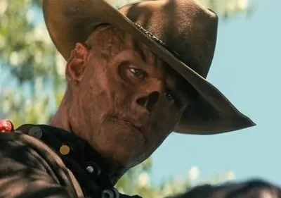 Close-up de um homem com chapéu de cowboy e rosto gravemente desfigurado, cicatrizado, com um olhar fixo e intenso sob céu azul.